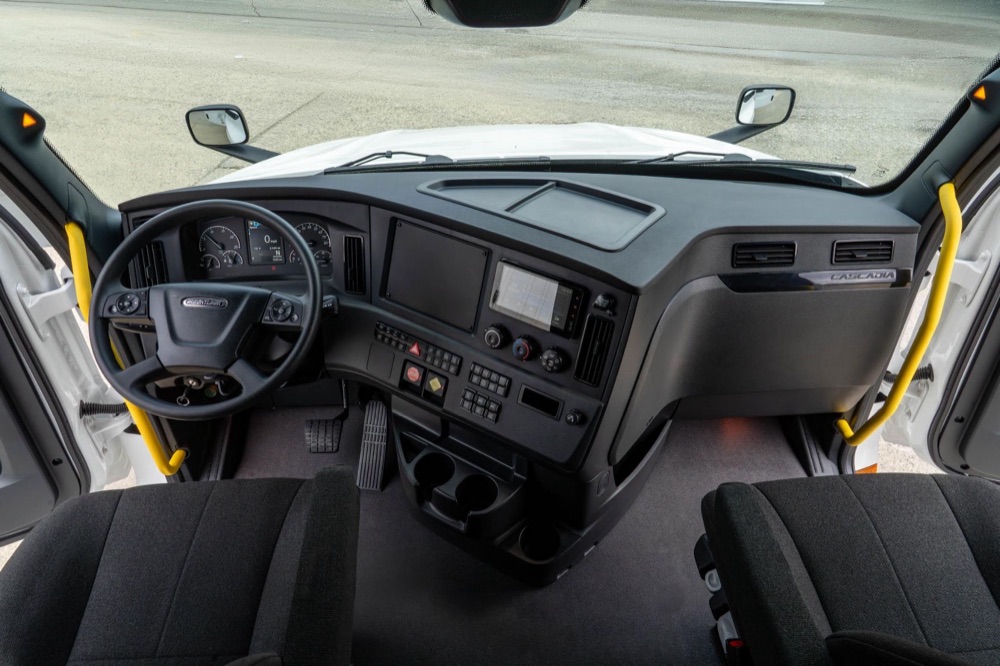 Inside the cab of a latest model Freightliner Cascadia: steering wheel, digital gauge cluster, touchscreen and dash controls.